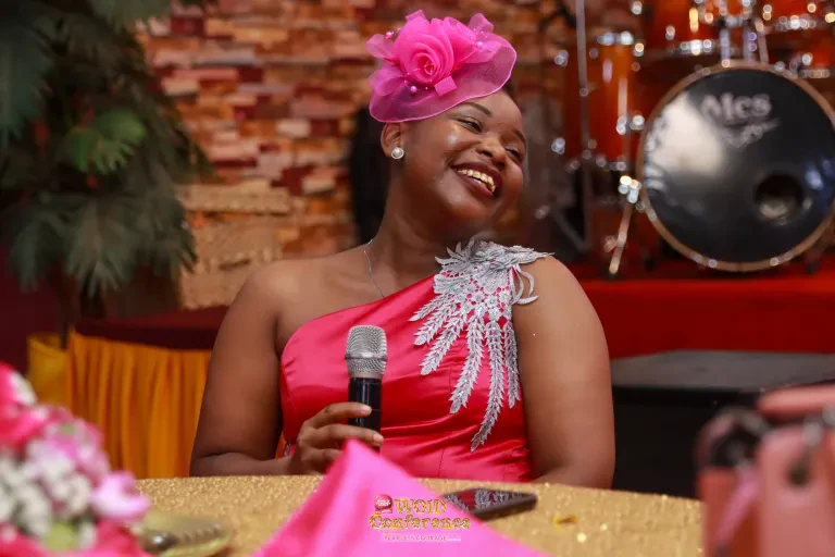 A woman in a pink fascinator laughs, microphone in hand, at the WOID@5 Dinner Night.