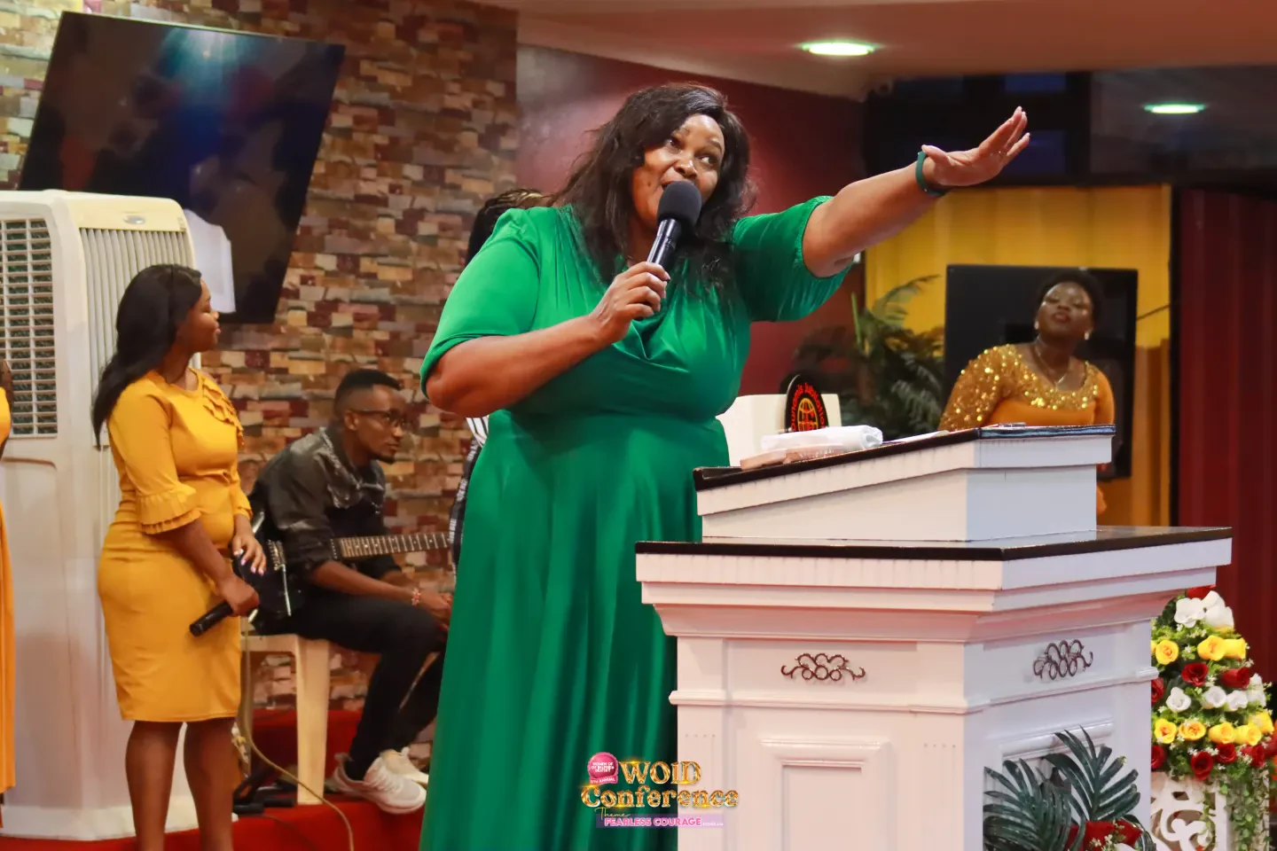 A speaker in a green dress preaches from the pulpit with her arm outstretched.