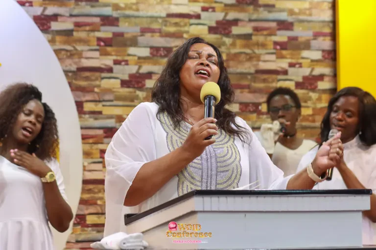 A woman in white leads worship at the pulpit, eyes closed, with singers behind her.