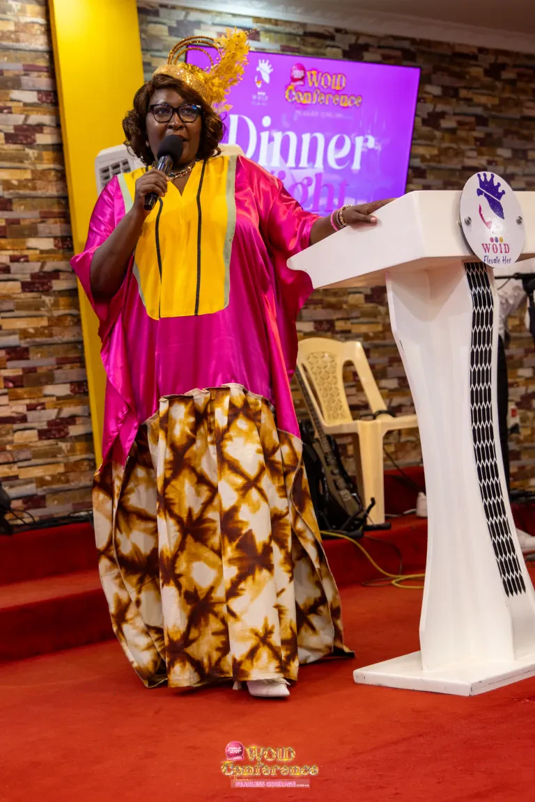 Rev. Pauline Mwarabu, in pink and gold with a gold fascinator, speaks at the WOID@5 Dinner Night.